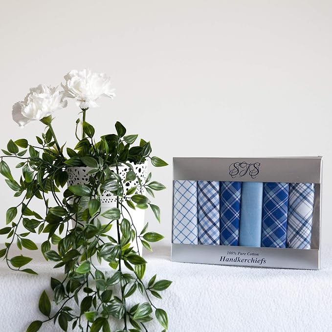 A boxed set of six blue patterned handkerchiefs sits on a white surface beside a white vase with green leaves and white flowers against a plain white wall.
