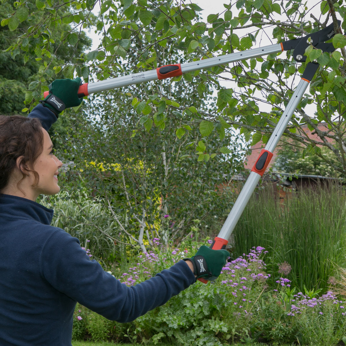 A woman in a navy fleece and green gloves uses a Telescopic garden loppers with bypass cutting action for pruning trees and bushes to trim green bushes in a garden.