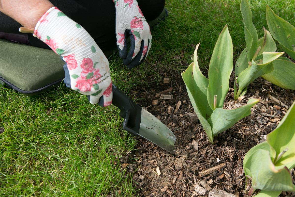 A close-up of a person kneeling in a garden, using a small hand trowel to dig around tulip plants. The person is wearing white gardening gloves with a pink rose pattern, and the scene is set on green grass and mulch.