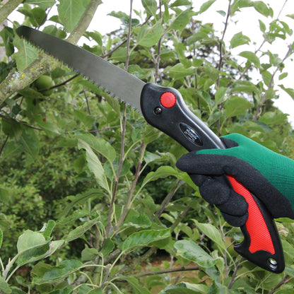 A hand wearing a green and black gardening glove holds a folding hand saw with a long silver blade and red accents, cutting through branches in a leafy garden.