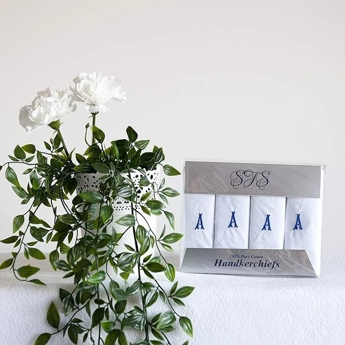 A set of four white handkerchiefs with blue embroidered letters A, A, A, and A, packaged in a silver box next to a white vase with green leaves and white flowers on a white surface against a white wall.