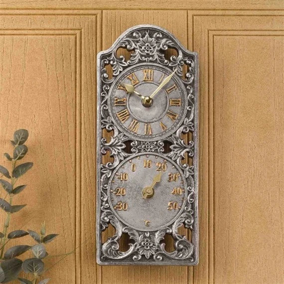 An ornate vintage-style wall clock with two circular dials, mounted on a light wood-paneled wall. The clock has an intricate silver-toned metal frame with Roman numerals on the upper dial and a temperature scale on the lower dial, with gold-toned hands.