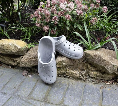 A pair of light gray Croc-style clogs placed on a stone garden border with flowers and greenery in the background.