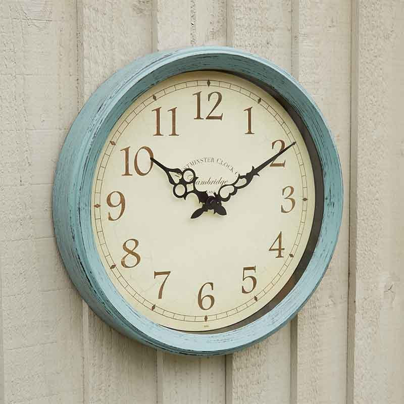 A round wall clock with a distressed light blue painted frame and a cream-colored dial, mounted on a light beige wooden wall. The clock features black Arabic numerals and ornate black hands.