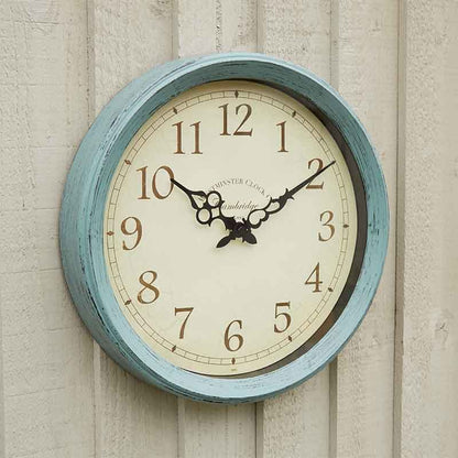 A round wall clock with a distressed light blue painted frame and a cream-colored dial, mounted on a light beige wooden wall. The clock features black Arabic numerals and ornate black hands.