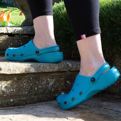 A person wearing bright turquoise blue Croc-style clogs outdoors on stone steps, with black leggings visible and grass in the background.