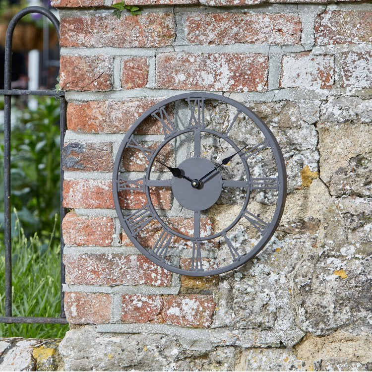 A round metal wall clock with Roman numerals and black hands, mounted on a weathered brick and stone wall outdoors.