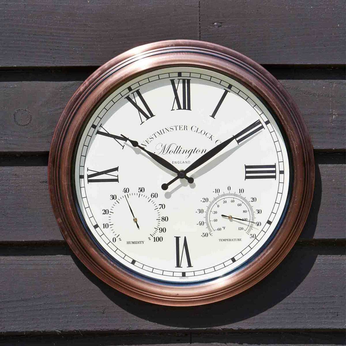 A round wall clock with a copper-toned frame and an off-white dial, mounted on dark wooden siding. The clock face features black Roman numerals, black hands, and two smaller sub-dials displaying temperature and humidity.