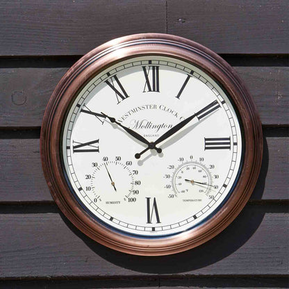 A round wall clock with a copper-toned frame and an off-white dial, mounted on dark wooden siding. The clock face features black Roman numerals, black hands, and two smaller sub-dials displaying temperature and humidity.