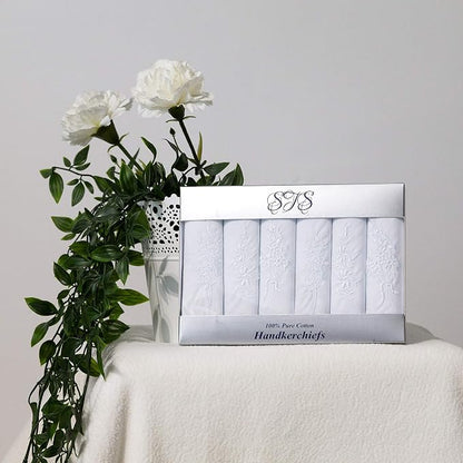 A boxed set of white handkerchiefs displayed on a draped white surface, with white flowers and green leaves beside the package against a light gray background.
