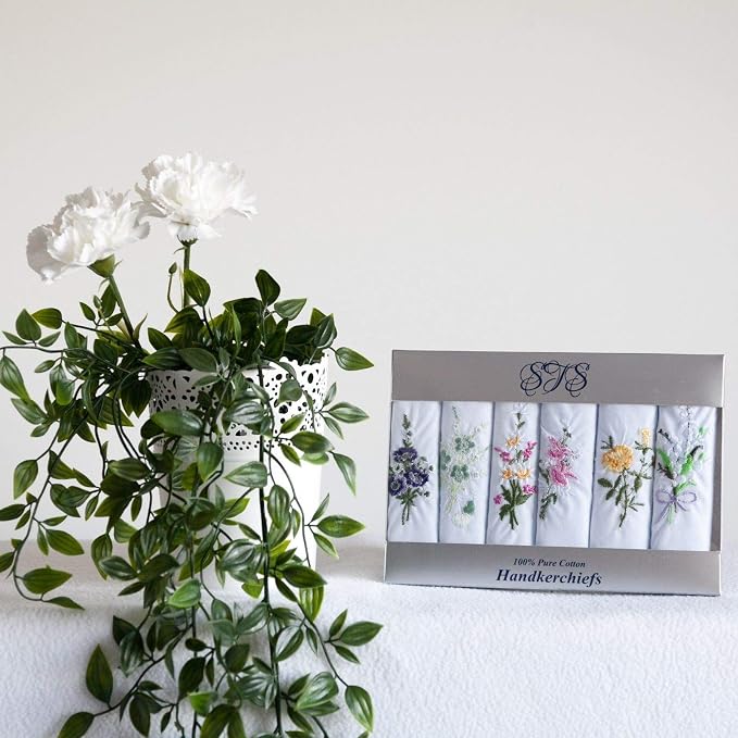 A boxed set of six folded white cotton handkerchiefs with floral embroidery, displayed next to a white vase with green leaves and white flowers against a plain light background.