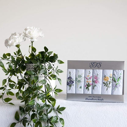 A boxed set of six folded white cotton handkerchiefs with floral embroidery, displayed next to a white vase with green leaves and white flowers against a plain light background.