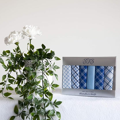 A boxed set of six blue patterned handkerchiefs sits on a white surface beside a white vase with green leaves and white flowers against a plain white wall.