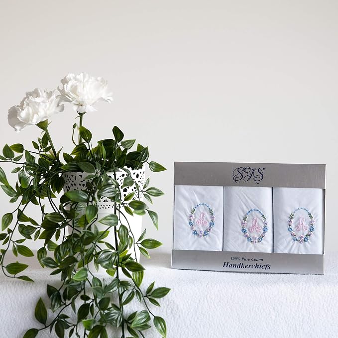 A boxed set of three white handkerchiefs with embroidered pastel monograms and floral details, displayed beside a white vase with green leaves and white flowers on a light background.