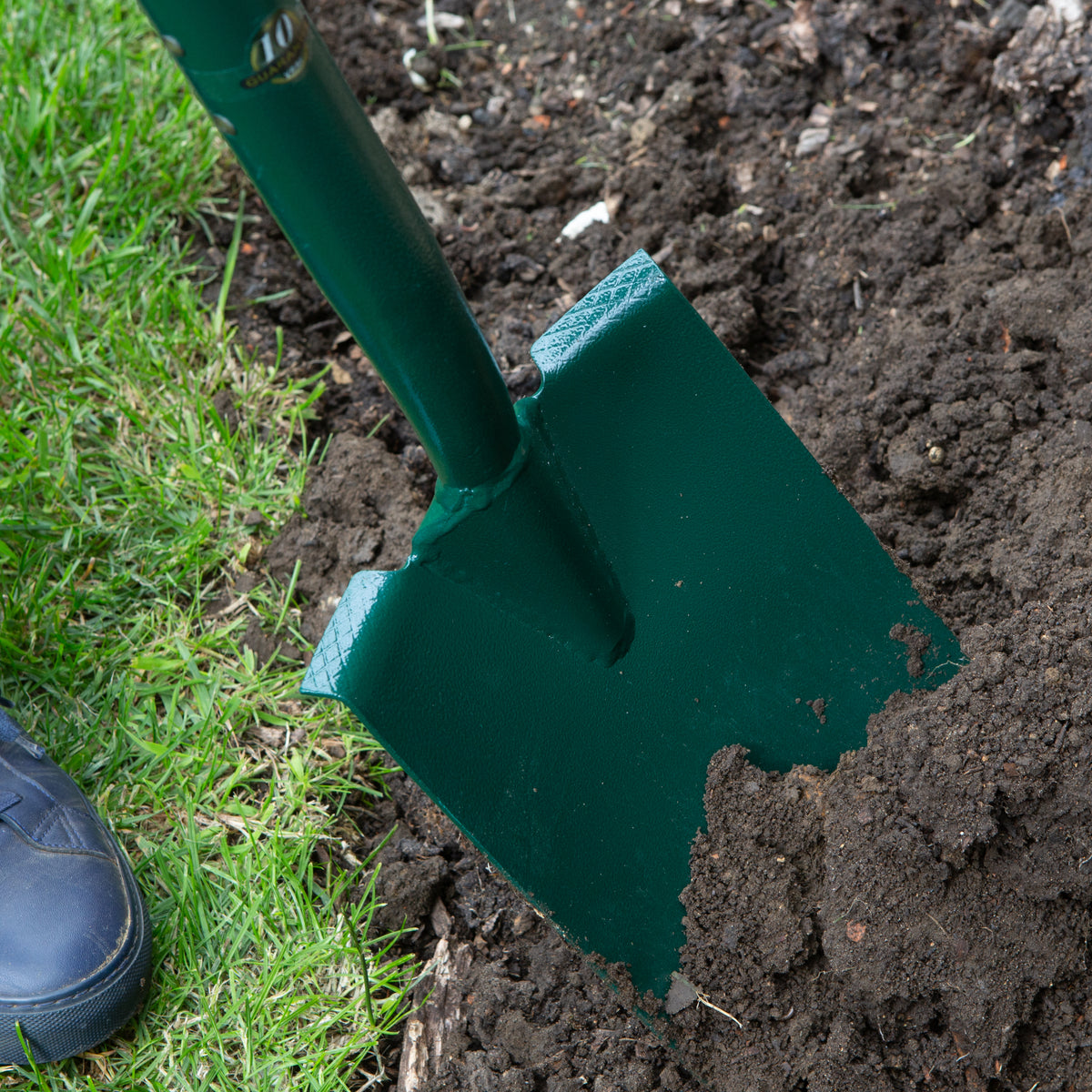 A dark green metal garden spade with a matching handle, shown digging into dark soil next to green grass, with a black shoe visible in the lower left corner.