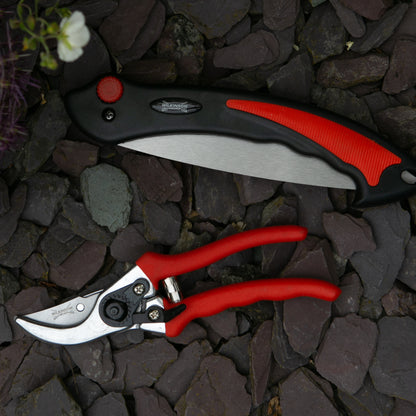 A pruning knife and matching pruning shears laid on dark gray stones. The pruning knife has a black and red handle with a silver blade, and the shears have red handles with silver metal blades and pivot.