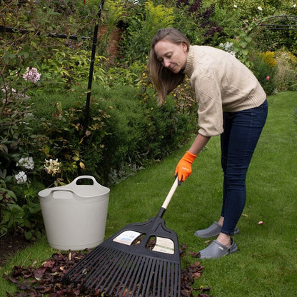 A woman in a beige sweater, dark jeans, and orange gardening gloves uses a black leaf rake to gather leaves on a green lawn, with a white bucket nearby in a garden setting.