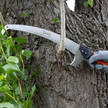 A close-up of a person's gloved hand using a pruning saw with a curved silver metal blade to cut a thin tree branch protruding from rough brown tree bark outdoors.
