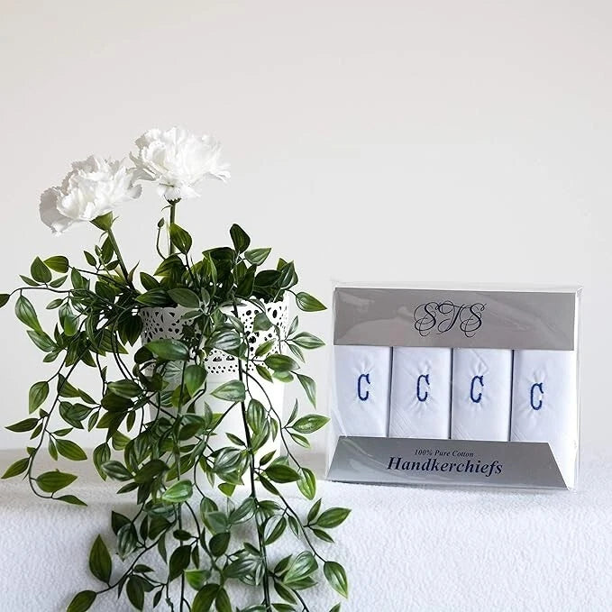 A boxed set of four white handkerchiefs with blue embroidered letters C displayed next to a white floral arrangement on a white surface against a white background.