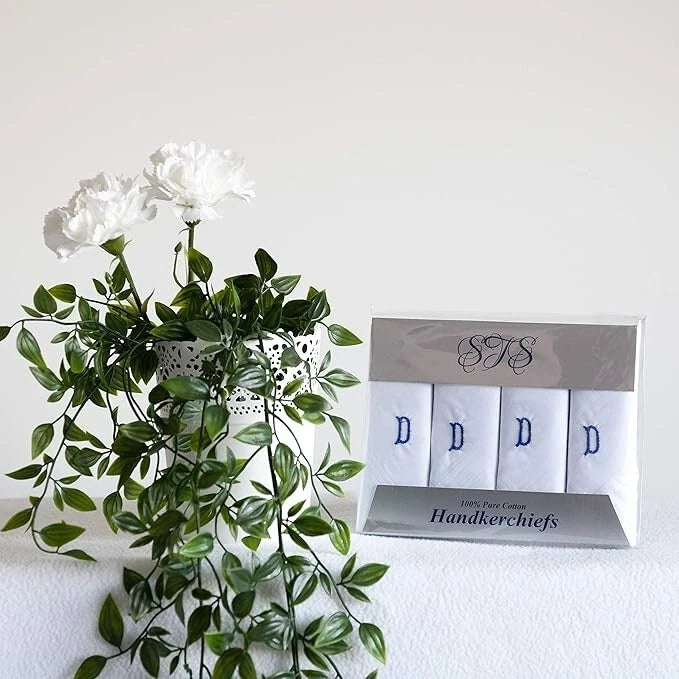 A boxed set of four white handkerchiefs with blue embroidered letters D displayed next to a white vase with green leaves and white flowers on a white surface against a white wall.