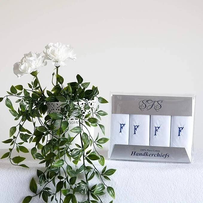 A boxed set of four white handkerchiefs with blue embroidered letters F displayed next to a white flower arrangement on a white surface against a white wall.