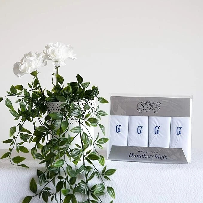 A set of four white handkerchiefs with blue embroidered monogram letters G, displayed in a clear plastic case next to a white flower arrangement on a white surface against a white background.