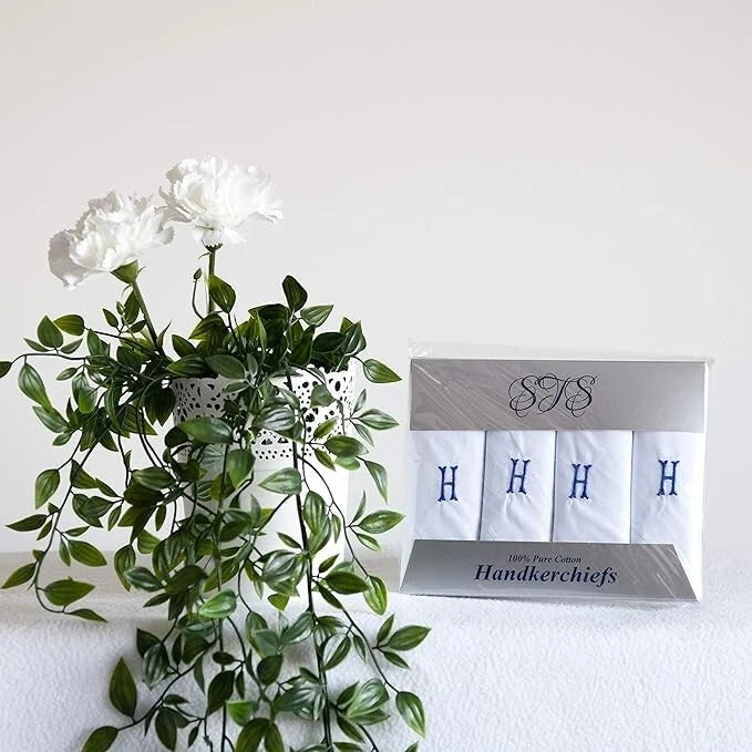 A boxed set of four white handkerchiefs with blue embroidered letters H on each square, displayed next to a white vase with green trailing leaves and white flowers against a plain white background.