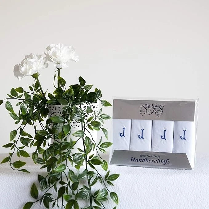 A boxed set of four white handkerchiefs with blue embroidered monogram letters, displayed next to a green artificial plant on a white surface against a white wall.