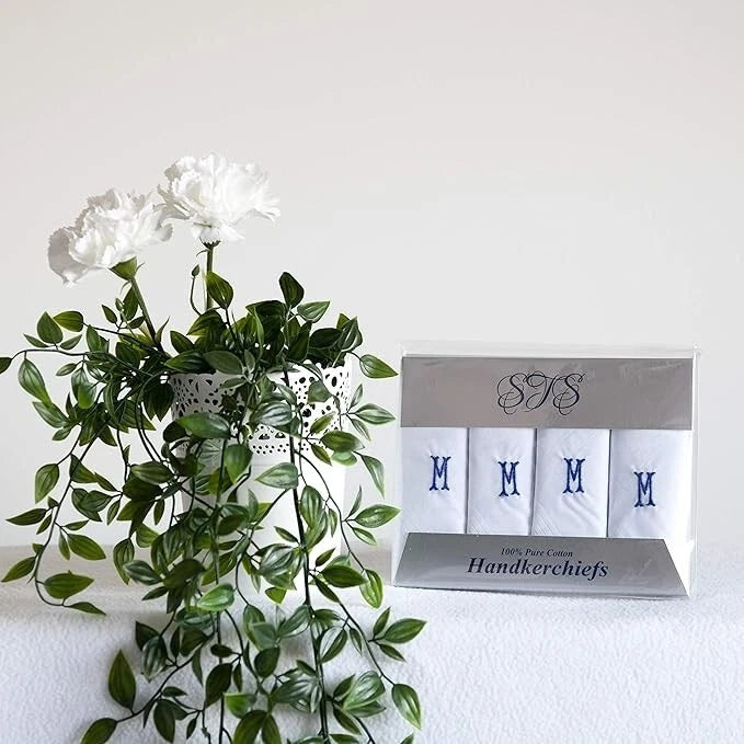 A boxed set of four white handkerchiefs with blue monogram letters 'M' sits beside a white vase with green trailing leaves and white flowers against a plain white wall.