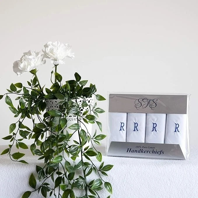 A boxed set of four white handkerchiefs with blue monogram letters R, displayed next to a green artificial plant on a white surface against a white wall.