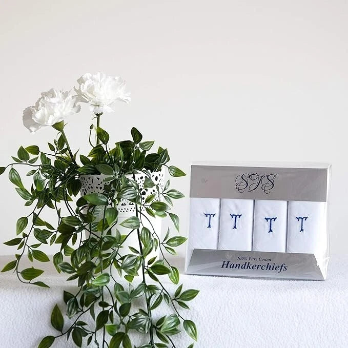 A boxed set of four white handkerchiefs with blue embroidered letters, displayed next to a white artificial flower arrangement on a white surface against a white wall.