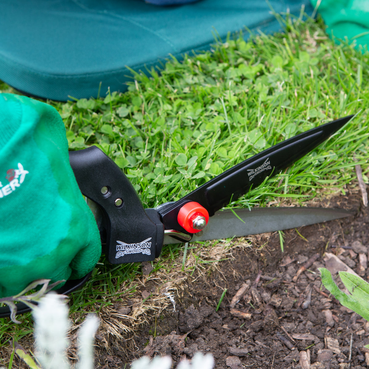A close-up of  Handheld grass shears with sharp blades for maintaining neat lawn borders with black blades and handles, a red adjustment knob, and a green fabric sheath, shown on grass and soil outdoors.