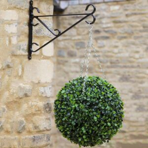 A spherical green artificial topiary tree hanging from a black metal wall bracket against a beige stone wall.