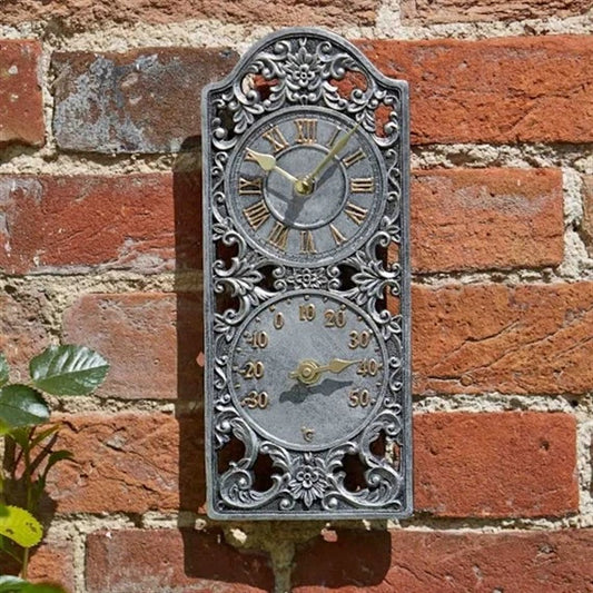 An ornate vintage-style metal wall clock with a decorative filigree frame, featuring a Roman numeral face on top and a temperature gauge on the bottom, mounted on a red brick wall.