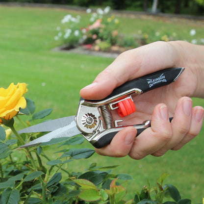 A hand holding a Wilton brand pruner with a black and silver handle and a red release button, cutting a yellow rose in a green garden.