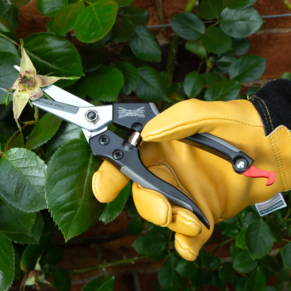 Hand wearing a yellow glove holding Straight garden snips with hard chrome-plated SK5 Japanese steel blades against green leaves
