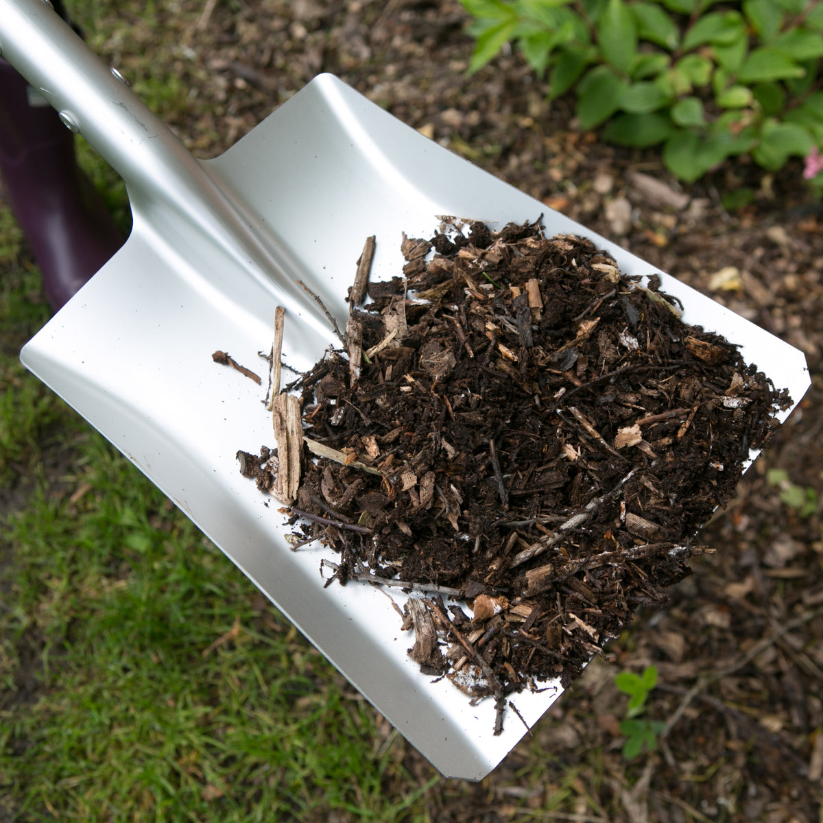 A white garden shovel filled with dark brown mulch or compost, photographed outdoors on grass and mulch with a pair of purple boots partially visible at the left edge.