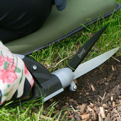 Close-up of a person using garden shears with a floral-patterned glove to cut grass and soil outdoors.